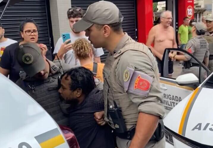 Policiais levam o motoboy Éverton à viatura policial. Ao fundo, à direita, o senhor apontado como agressor conversa com uma policial. Foto: reprodução/Instagram (@nietzsche4speed)