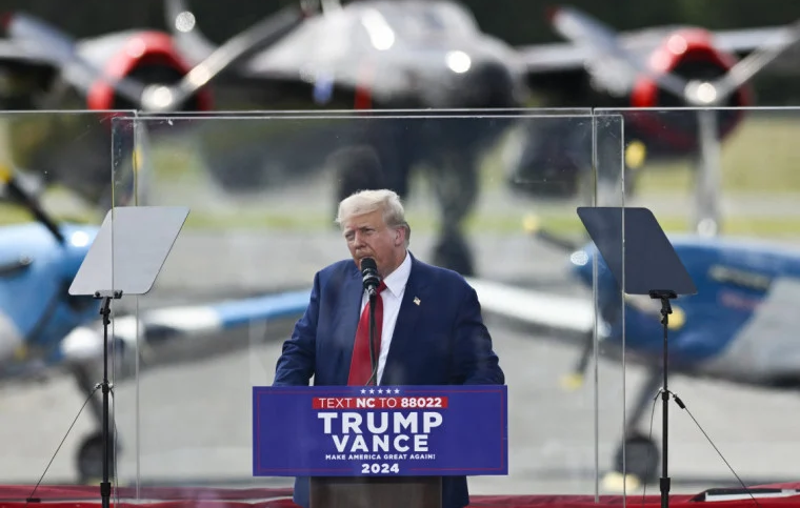 Trump discursando atrás de blindagem de vidro após atentado, reforçando a ideia de segurança como símbolo político.