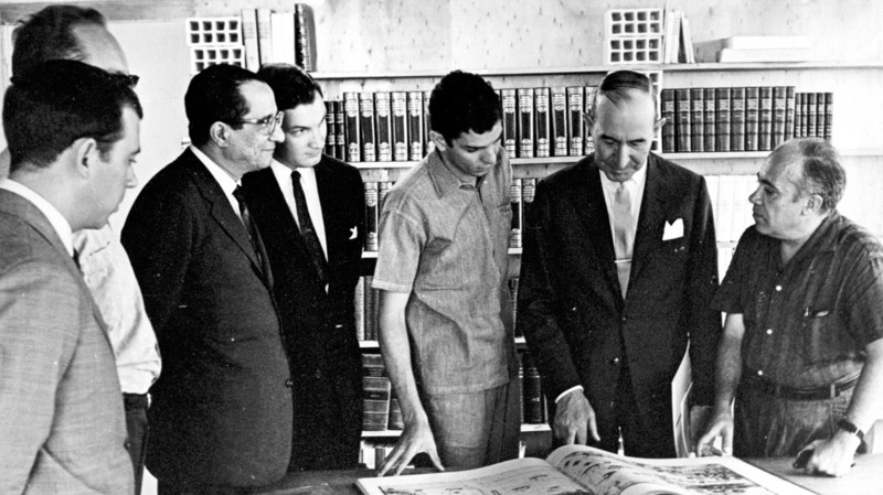 Na foto da década de 1960, Eudoro de Sousa (o último da esquerda para a direita) mostra o espaço do Centro de Estudos Clássicos para uma comitiva de visitantes.&nbsp; Ao fundo pode-se ver parte do acervo do setor de referências da biblioteca do CEC que, na época, ocupava o subsolo do prédio da reitoria. &nbsp;